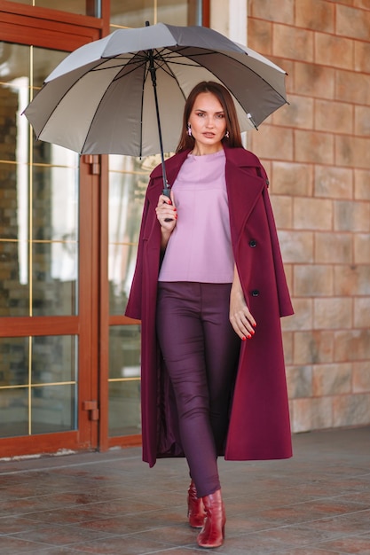 A model girl showing fashionable outerwear with an umbrella in her hand on the street on the veranda of a summer cafe Clothes for the showroom