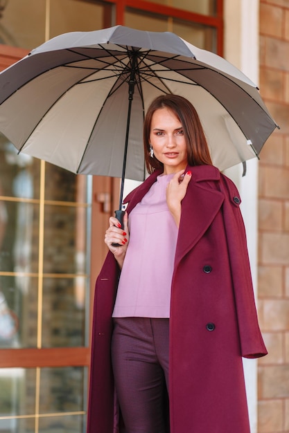 A model girl showing fashionable outerwear with an umbrella in her hand on the street on the veranda of a summer cafe Clothes for the showroom