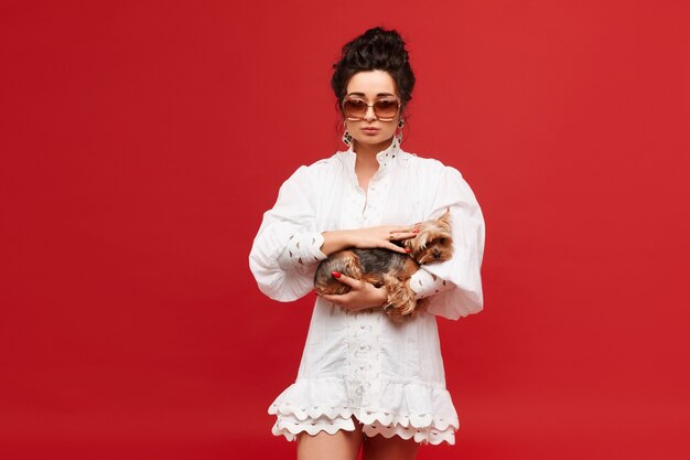 Model girl with curly black hair in fashionable sunglasses and white dress keeps a yorkshire terrier...