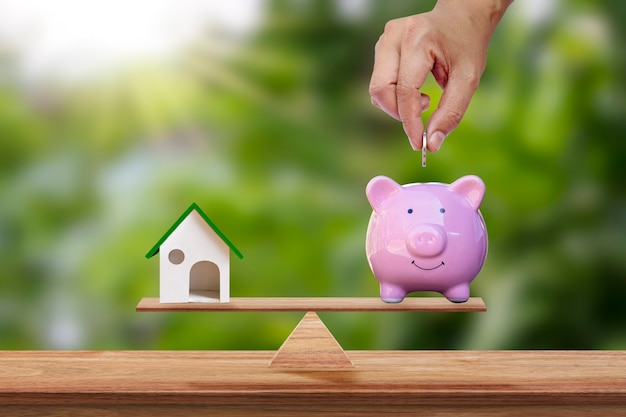 A model house and a hand holding a coin in a piggy bank that is placed on the scale