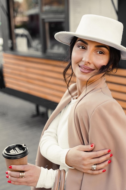 Model met koffiekopje in de buurt van coffeeshop. Meisje droeg witte jurk, jas en hoed poseren in de buurt van café.