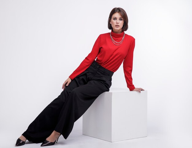 Model in pants red top necklace on white background Bob haircut Studio Shot Model sits on cube