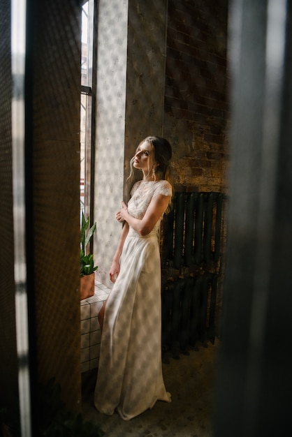 Model poseren in een witte lange trouwjurk binnenshuis door het raam.