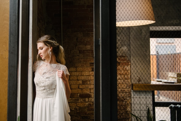 Model poseren in een witte lange trouwjurk binnenshuis door het raam.