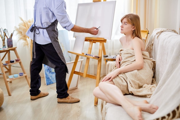 Model posing for portrait in studio, cropped male in apron drawing her portrait on canvas