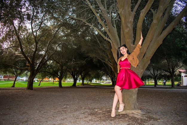 Модель в красном платье позирует рядом с деревом в парке.