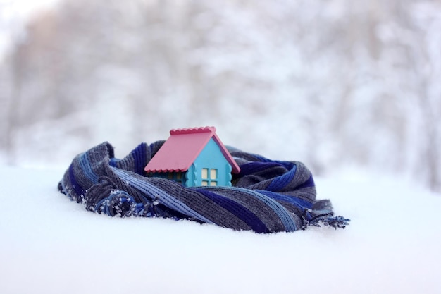 A model of a rustic house wrapped in a warm scarf on a blurry winter background A toy cottage in the snow