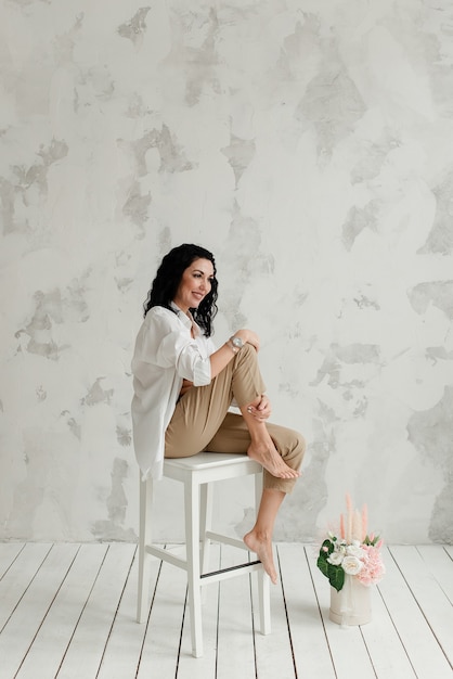 Model shooting in the studio a brunette girl sits on a white chair with a bouquet of flowers