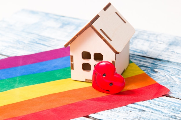 Макет небольшого деревянного домика и сердечко стоит на флаге цветов радуги, синей деревянной поверхности.