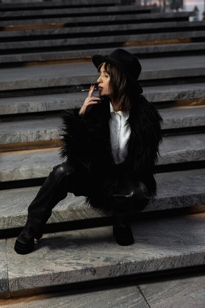 Model smokes.Outdoor fashion portrait of young beautiful girl wearing trendy black faux fur coat.