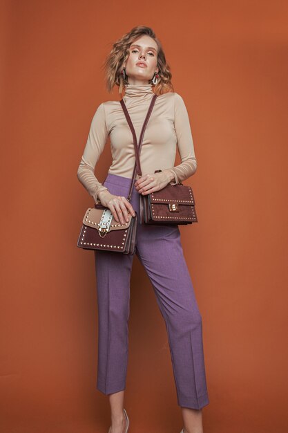 Model standing in beige polo-neck and purple trousers and holding two bags.