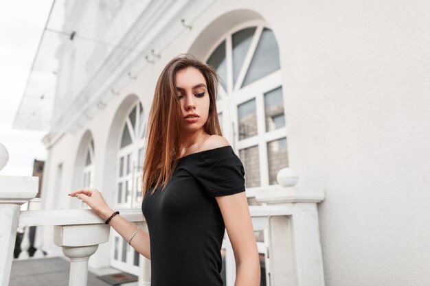 Model of a stylish young woman with long brown hair in a sexy black dress posing on the street near a vintage white building. Attractive fashionable girl walks around the city on an autumn day.