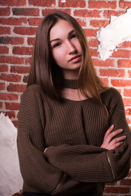 Model test of attractive brunette model with natural makeup, wears oversize sweater, posing on a brick wall background