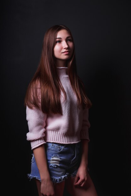 Model test of beautiful brunette model with long hair, wears pink sweater,blue jeans shorts, posing on a black background