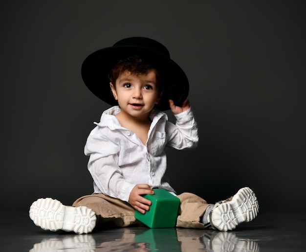 Model toddler baby wearing casual clothes shoes and a hat\
holding a toy building block isolated portrait isolated shot on\
gray studio background fashionable outfit for children\'s\
advertising