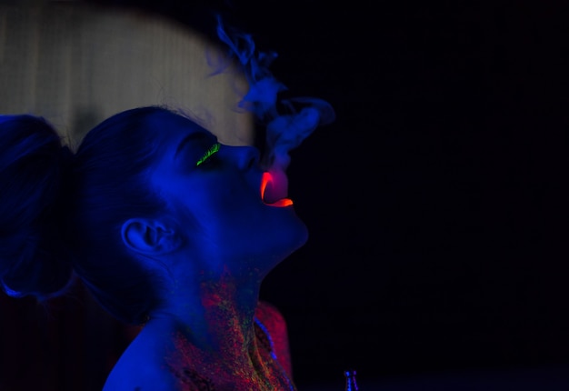 model in uv neon light with fluorescent glowing Body Art makeup She blows smoke from the hookah