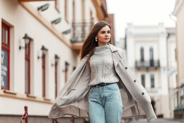 model van een jonge vrouw in een trenchcoat en een gebreide vintage trui loopt over straat