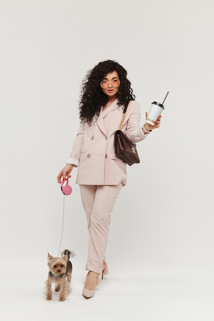 La donna di modello nella tenuta dell'attrezzatura alla moda porta via la tazza di caffè e cammina con il piccolo yorkie terrier