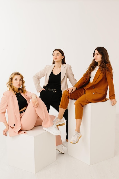Model women dressed in fashionable pastel blazers and pants with stylish sneakers posing on a cube on a white background in the studio