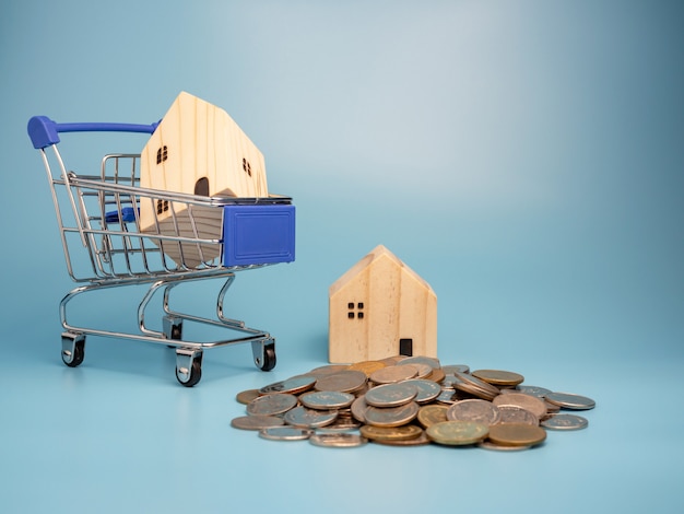 Photo a model wooden house on shopping cart with a pile of coins on blue