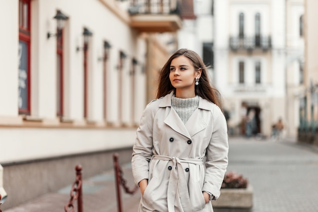 model of a young woman in a trench coat and a knitted vintage sweater walks down the street