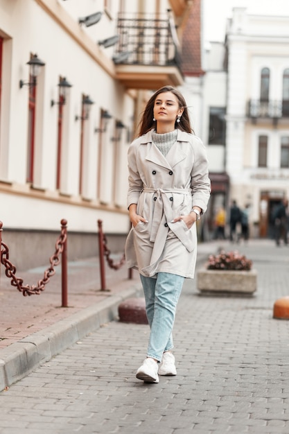 model of a young woman in a trench coat and a knitted vintage sweater walks down the street