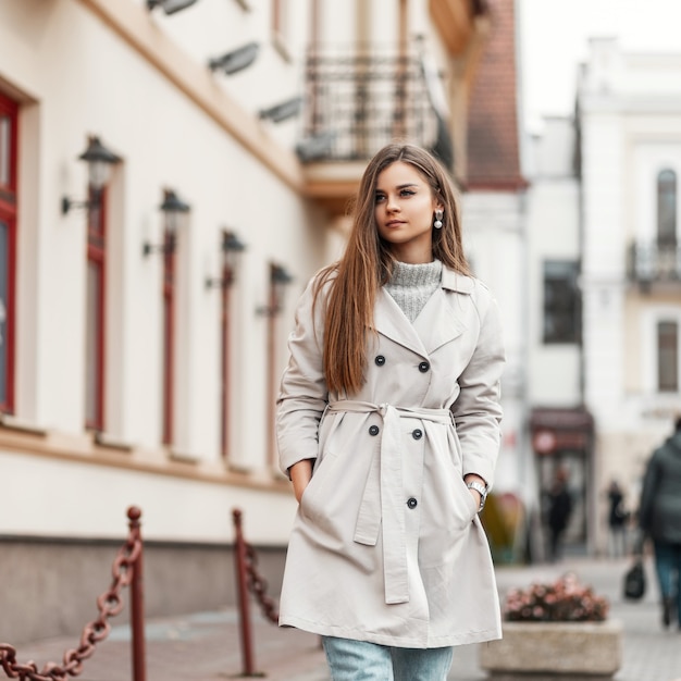 model of a young woman in a trench coat and a knitted vintage sweater walks down the street