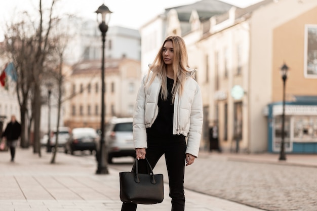 model of a young woman in a white jacket and a leather trendy bag stands on the street