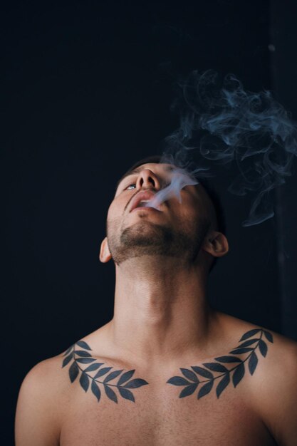 Modelling snapshots pensive focused tanned attractive handsome\
naked man tipping his head back smoking posing isolated in black\
studio background fashion offer copy space for ad closeup