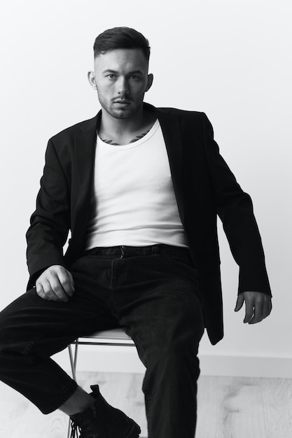 Моделирование снимков Серьезный самоуверенный привлекательный красивый мужчина в черной куртке сидит на стуле и позирует на белом фоне студии Черно-белая концепция Копирование пространства