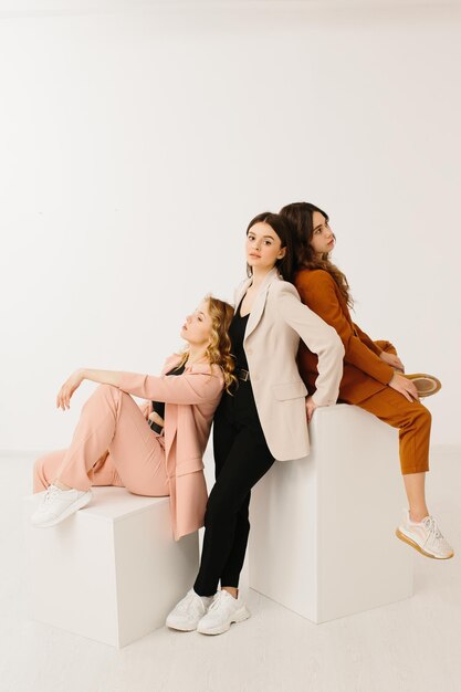 Modelvrouwen gekleed in modieuze pastelkleurige blazers en broeken met stijlvolle sneakers poseren op een kubus op een witte achtergrond in de studio