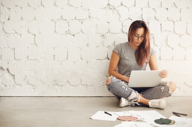 Modeontwerper student met laptop ontdekt ideeën over haar nieuwe collectie omringd door textiel en schetsen zittend op de vloer in haar studio