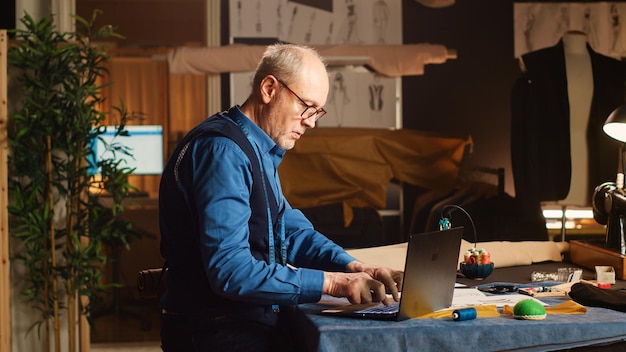 Modeontwerper werkt aan kledingontwerp met schetsen, gebruikt een laptop om maten en op maat gemaakte stoffen te bekijken. Mannelijke couturier die luxe kleding maakt in een klein zakelijk atelier.