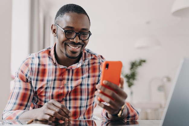Modern african american man in glasses uses mobile apps holding smartphone sitting at desk smiling