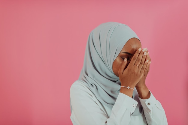 Modern african muslim woman makes traditional prayer to god,\
keeps hands in praying gesture, wears traditional white clothes,\
has serious facial expression, isolated over plastic pink\
background. high