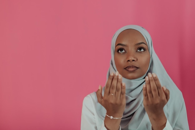 Modern African Muslim woman makes traditional prayer to God, keeps hands in praying gesture, wears traditional white clothes, has serious facial expression, isolated over plastic pink background. High