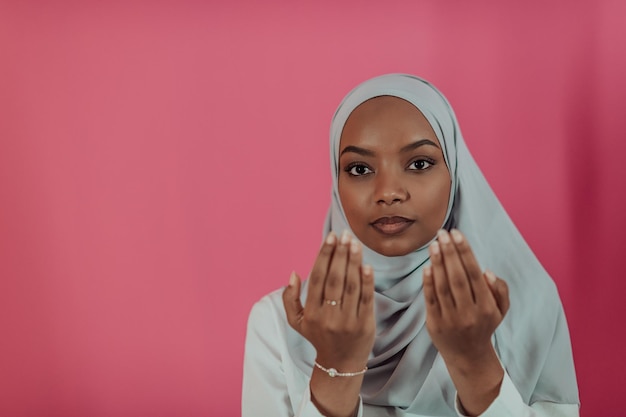 Modern African Muslim woman makes traditional prayer to God, keeps hands in praying gesture, wears traditional white clothes, has serious facial expression, isolated over plastic pink background. High