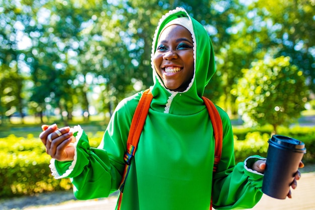 Modern afro woman in green hijab with bright make up and piercing nose drinking coffee outdoor and communicating with her friend