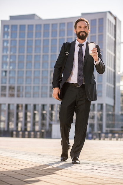 Modern architecture. Full length of a joyful handsome businessman drinking coffee outdoors while going to work