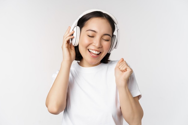 Ragazza asiatica moderna che balla musica d'ascolto con le cuffie che sorridono felice in piedi in maglietta sopra priorità bassa bianca