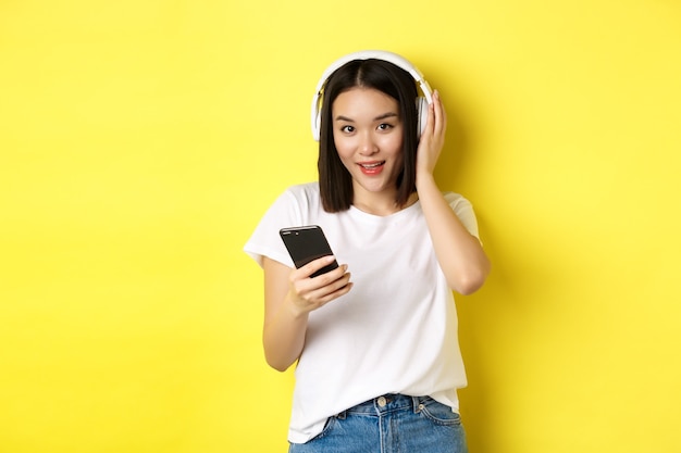 Modern asian woman listening music in wireless headphones, reading smartphone screen and smiling, standing in white t-shirt over yellow.