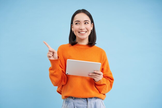 Modern asian woman with tablet pointing at empty space for promo offer banner smiling and looking person