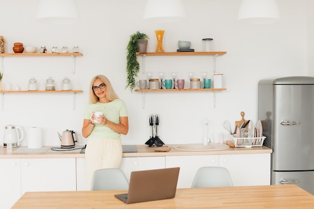 Modern attractive middle-aged woman drinking coffee and working on laptop in kitchen at home