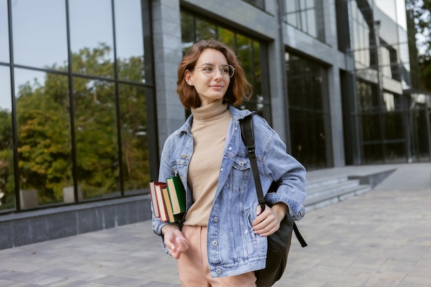 Modern attractive woman student in stylish clothes with stack of books and backpack outdoors