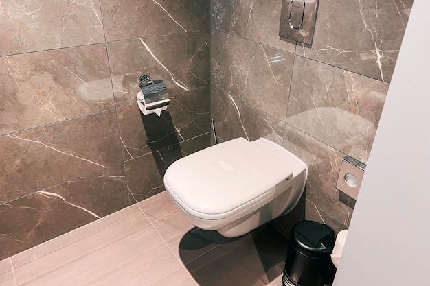 A modern bathroom with a ceramic toilet in white The toilet is closed and has a seat and a flush The bathroom has a wall and a floor with tiles