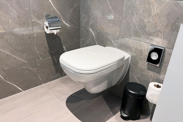 A modern bathroom with a white ceramic toilet The toilet is closed and has a stylish seat and a flush The bathroom has a wall and a floor made of tiles