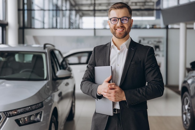 modern bearded man in glasses and suit vehicle sales consultant using laptop inside car dealership