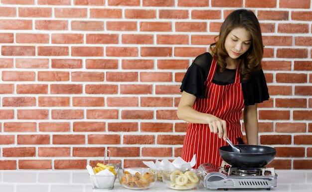 Modern beautiful Asian housewife in red striped apron smiling as enjoy lifestyle of cooking fried food on table near brick wall in kitchen by putting onion rings from glass bowl into black pan