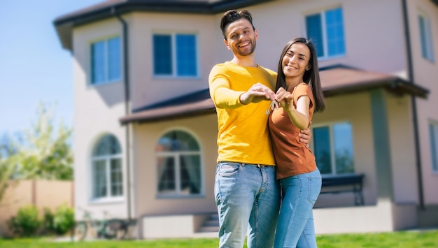 Modern beautiful excited young couple in love standing in front of the new big house with the keys in hands and hugging while they celebrating this buying