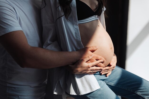 modern beautiful pregnant woman in jeans and shirt with man hugging her tummy with hands in beautiful sunlight Concept of pregnancy motherhood preparation and waiting